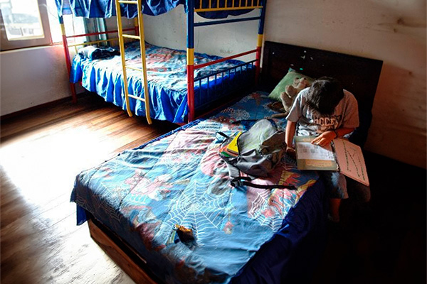 La imagen muestra un niño en un cuarto sentado en la cama con varios cuadernos.